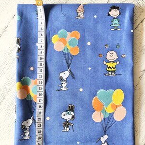 May include: Blue fabric with a white polka dot pattern featuring various cartoon characters from the Peanuts comic strip. The characters include Snoopy, Charlie Brown, and Lucy. Snoopy is depicted holding a bunch of colorful balloons, while Charlie Brown is juggling balls and Lucy is standing with her arms crossed.