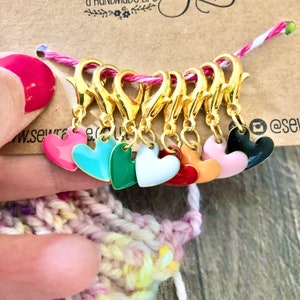 May include: Set of eight gold heart-shaped stitch markers with different colored enamel finishes. The colors include pink, turquoise, green, white, red, orange, peach, and black.