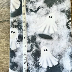 May include: White fabric with a black and white Halloween print featuring ghosts, bats, and spiders. The fabric is measured with a ruler in inches.