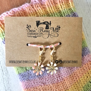 May include: Two gold stitch markers with flower charms. The charms are white with pink centres and gold accents. The stitch markers are on a brown card with the text "Sew Ray Me a handmade life" and the website address "www.sewrayme.co.uk".