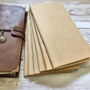 May include: A brown leather journal with a compass charm and a stack of blank kraft paper notebooks. The journal has decorative corner accents. The notebooks are stapled together and ready for writing.