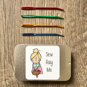 May include: A silver metal tin with a white label that says "Sew Ray Me". The label has a cartoon illustration of a girl with blonde hair holding a basket of yarn. The tin contains four colourful sewing needles.