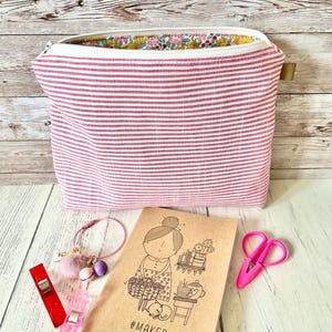 May include: A pink and white striped zippered pouch with a floral interior lining. A notebook with a drawing of a woman and the text "#MAKER Project Notes" is next to the pouch, along with pink scissors, a bracelet, and sewing clips.