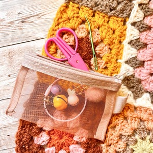 May include: A selection of crochet tools and accessories, including pink scissors, a green needle, and a mesh pouch with decorative charms. The items are placed on a colourful, handmade granny square blanket in shades of brown, yellow, and pink.