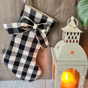 May include: A black and white checked Christmas stocking with a bow, hanging on a wooden surface. A white lantern with a lit candle is next to it, creating a festive holiday scene.