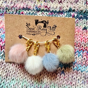 Peut inclure: Cinq pompons en fausse fourrure de couleurs différentes attachés à des marqueurs de points en métal doré. Les marqueurs de points sont sur une carte brune avec le texte "Sew Ray Me a handmade life".