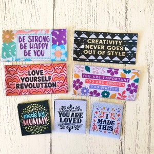 Könnte beinhalten: Ein Set aus zehn bunten Stoff-Etiketten mit inspirierenden Botschaften. Die Etiketten zeigen verschiedene Designs, darunter Herzen, Blumen und geometrische Muster. Die Botschaften lauten: "Sei stark Sei glücklich Sei du", "Liebe dich selbst Revolution", "Kreativität kommt nie aus der Mode", "Du bist wichtig Du bist genug Du zählst", "Gemacht von Mama", "Du bist geliebt", und "Ich habe das gemacht".