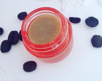 May include: A small, clear jar with a red lid filled with a light brown lip balm. The jar is sitting on a white surface with coffee beans scattered around it.