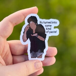 May include: A sticker featuring a cartoon illustration of a person with dark hair and a black shirt. The person is holding a small object in their hand. The sticker has the text "Its turned into some kind of paste!" printed on it.