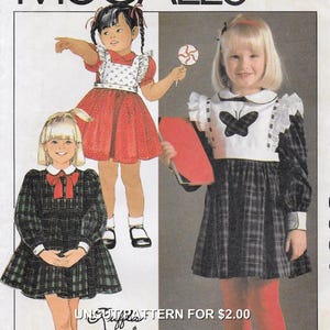 May include: Vintage sewing pattern featuring illustrations of three little girls in dresses. The dresses include a red dotted dress with a white apron, a plaid dress with a red bow, and a plaid dress with a butterfly design. The text "UN-CUT PATTERN FOR $2.00" is visible.