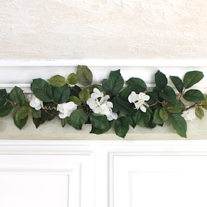 May include: A decorative garland featuring white flowers, dark berries, and green leaves. The garland is arranged on a white surface, creating a natural and elegant aesthetic. Perfect for adding a touch of nature to any space.