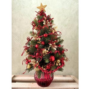 May include: A small artificial Christmas tree decorated with red and gold ornaments, red beaded garlands, and gold ribbons. The tree is topped with a gold star and is set in a ribbed, translucent red glass vase with a rounded, bulbous base.