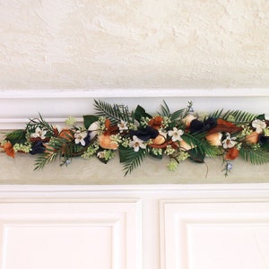 May include: A decorative floral garland featuring a mix of faux flowers, leaves, and berries. The arrangement includes white, orange, and navy blue elements, with green foliage and brown accents. It is displayed above white cabinets.