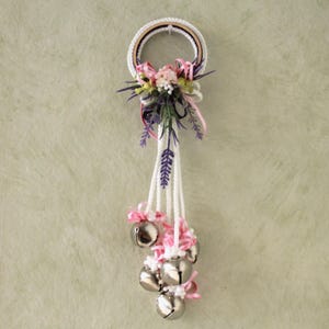 May include: A white crocheted wreath with pink and purple flowers and a string of silver bells hanging from it.