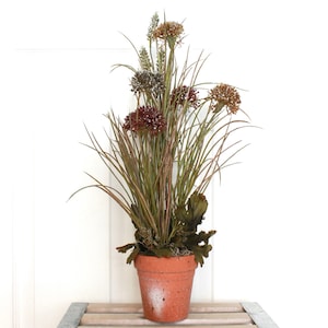May include: An artificial plant arrangement in a terracotta-colored pot. The plant features tall, thin green grass blades and several round, textured flower heads in shades of deep burgundy, brown, and muted green, creating a dense, upright display.