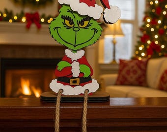 May include: A decorative wooden cutout of a green character with a mischievous expression, wearing a red and white Santa hat and suit. The figure has dangling legs made of jute rope ending in black curled shoes, designed to sit on a flat edge.