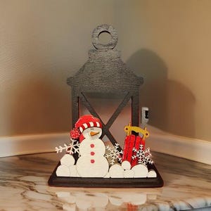 May include: A wooden Christmas lantern with a snowman and sled scene. The snowman wears a red and white hat and has a carrot nose. The sled is red with yellow accents. The lantern is dark gray with a decorative top.