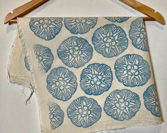May include: A piece of off-white fabric with a repeating pattern of blue floral designs. The fabric is hanging on a wooden hanger. The edges of the fabric are unfinished.