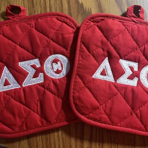 May include: Two red quilted pot holders with white embroidered Greek letters. Each pot holder has the letters ΔΣΘ. The pot holders have a loop for hanging and are on a wooden surface.