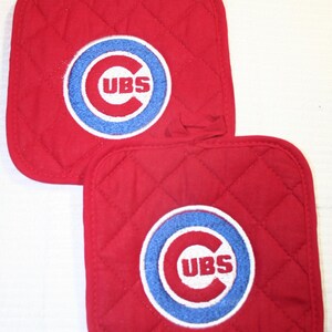 May include: Two red quilted pot holders with a white, blue, and red Chicago Cubs logo. The logo features a blue circle with the letters "UBS" in white. Each pot holder has a loop for hanging.