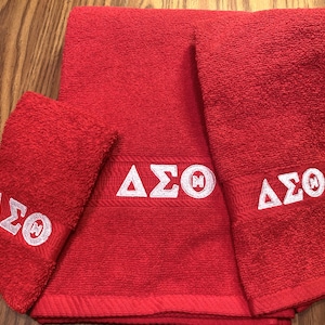May include: A set of three red bath towels. The towels are embroidered with the Greek letters ΔΣΘ in white. The towels are folded and arranged on a wooden surface.
