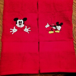 以下が含まれることがあります： ミッキーマウスの刺繍が施された赤いハンドタオル2枚。1枚は、ミッキーマウスが手を伸ばして上の方をのぞいている。もう1枚は、ミッキーマウスが腕を伸ばして歩いている。