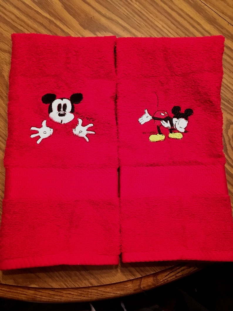 以下が含まれることがあります： ミッキーマウスの刺繍が施された赤いハンドタオル2枚。1枚は、ミッキーマウスが手を伸ばして上の方をのぞいている。もう1枚は、ミッキーマウスが腕を伸ばして歩いている。