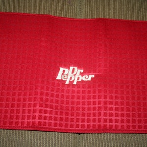 May include: A red rectangular kitchen towel with a textured, checkered pattern. The white embroidered "Dr Pepper" logo is centered on the towel. The towel is folded and appears to be made of a soft fabric.