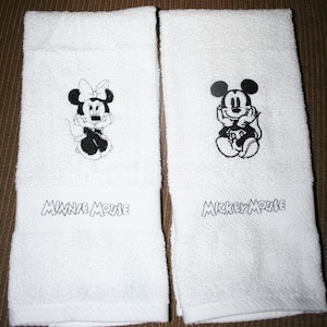 Peut inclure: Deux serviettes de toilette blanches avec des images brod&eacute;es noires de Mickey Mouse et Minnie Mouse. Les serviettes portent les noms "Mickey Mouse" et "Minnie Mouse" brod&eacute;s sous les images.