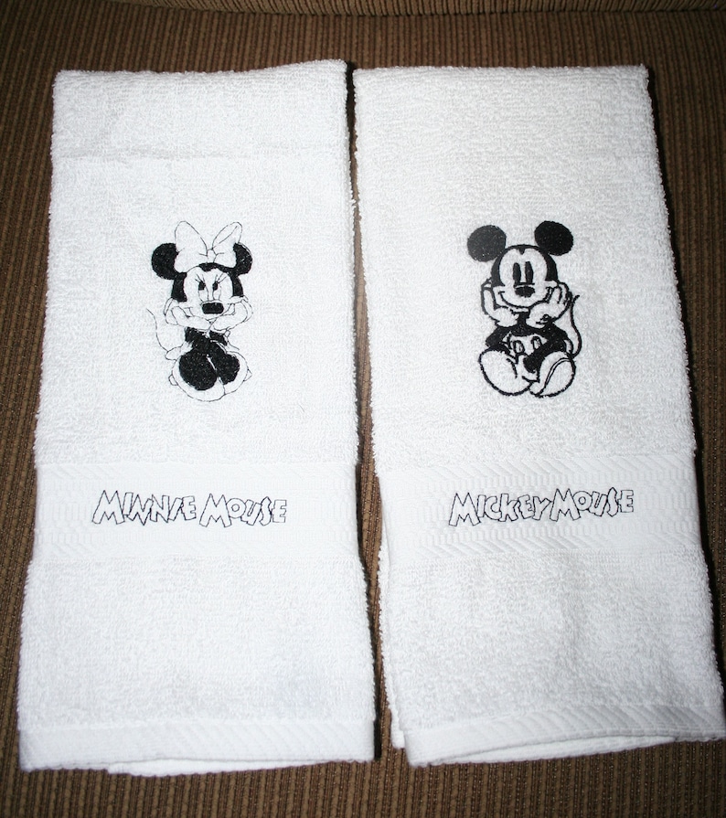 Peut inclure: Deux serviettes de toilette blanches avec des images brod&eacute;es noires de Mickey Mouse et Minnie Mouse. Les serviettes portent les noms "Mickey Mouse" et "Minnie Mouse" brod&eacute;s sous les images.