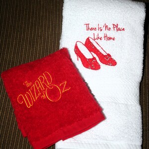 May include: Two embroidered towels, one red and one white. The red towel has "The Wizard of Oz" embroidered in gold thread. The white towel has "There is no place like home" and a pair of red ruby slippers embroidered in red thread.