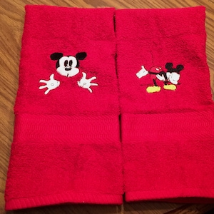 以下が含まれることがあります： 赤い手拭き2枚にミッキーマウスの刺繍が施されています。1枚は赤い背景からミッキーマウスが顔をのぞかせており、もう1枚はミッキーマウスが腕を広げて歩いている様子が描かれています。