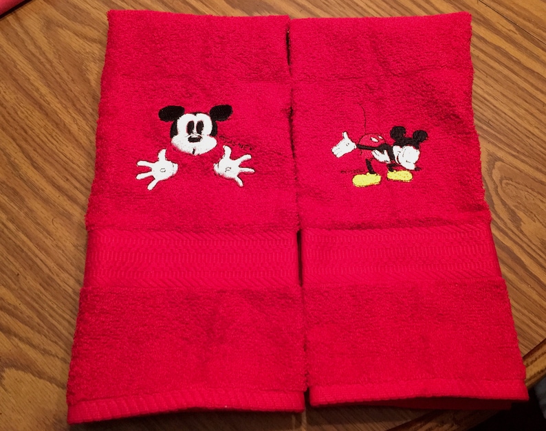 以下が含まれることがあります： 赤い手拭き2枚にミッキーマウスの刺繍が施されています。1枚は赤い背景からミッキーマウスが顔をのぞかせており、もう1枚はミッキーマウスが腕を広げて歩いている様子が描かれています。