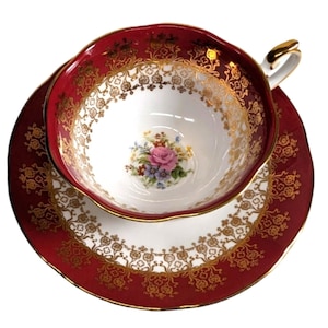 May include: A porcelain teacup and saucer set featuring a deep red border with intricate gold filigree patterns. The center of the cup displays a small floral bouquet with a pink rose and blue flowers. The cup has a gold-trimmed handle.