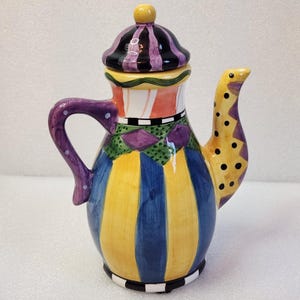 May include: A colorful ceramic teapot with a lid. The teapot features a purple handle, a yellow spout with black polka dots, and a body with vertical stripes in yellow and blue. The lid is black and purple striped.