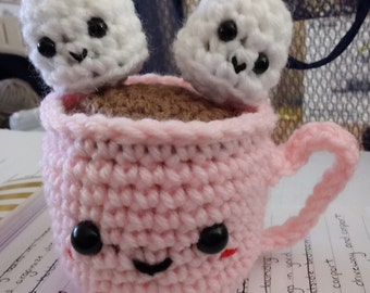 May include: A pink crocheted mug with a smiling face and two white crocheted marshmallows on top. The mug is filled with brown chocolate.