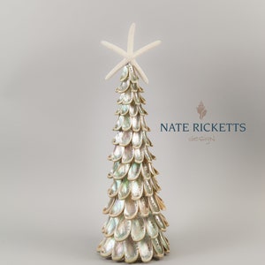 May include: A decorative Christmas tree made of iridescent shells, topped with a white starfish. The tree has a conical shape and is set against a light gray background. The tree is a unique coastal-themed holiday decoration.