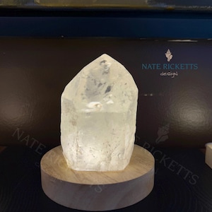 Op de afbeelding: Een helder, gefacetteerd kristal, van binnenuit verlicht, gemonteerd op een ronde, lichtgekleurde houten basis. Het kristal heeft een natuurlijke, ruwe textuur en een puntige top. De basis heeft een diameter van ongeveer 7.6 cm. De tekst "NATE RICKETTS design" is zichtbaar.