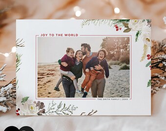 May include: A rectangular holiday photo card featuring a central photograph of a family of four on a beach. The card has a white border with watercolor illustrations of white flowers, green pine branches, and red berries. Text reads JOY TO THE WORLD and THE SMITH FAMILY | 20XX.