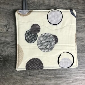 May include: A white quilted hot pad with a geometric pattern of black and gray circles. The circles have a textured fabric design.