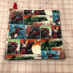 May include: A colorful fabric pot holder with a comic book pattern featuring superheroes like Hulk, Iron Man, Captain America, and Thor. The pot holder is made of cotton and has a red loop for hanging.