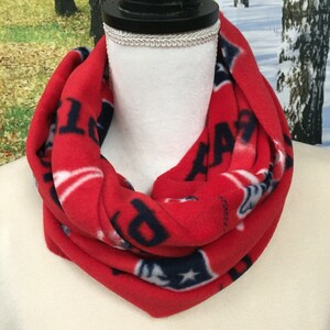 May include: A red fleece infinity scarf with a repeating pattern of white stars and navy blue text. The scarf is displayed on a white mannequin head. The scarf is a winter accessory.
