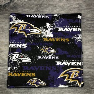 May include: Black and purple fabric with a repeated pattern of the Baltimore Ravens logo. The logo features a black raven with a white eye and a gold "B" on its chest. The words "Baltimore Ravens" are repeated throughout the fabric.