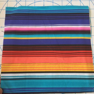 May include: A rectangular piece of fabric with a vibrant striped pattern. The design features horizontal bands of various colors, including turquoise, black, red, orange, yellow, and purple. A black stitch line runs across the fabric.