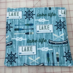 May include: A square, quilted fabric item with a light blue background and a lake-themed pattern. The design includes white rectangles with the word "LAKE" and arrows, along with images of oars, nautical wheels, knots, and canoes. The words "lake life" and "canoe time" are also present.