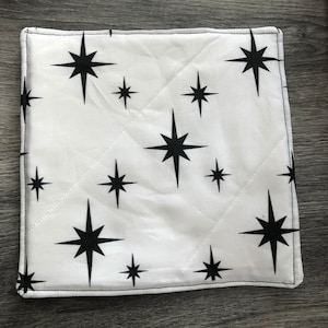 May include: White cotton kitchen potholder with a black starburst pattern. The potholder is quilted and has a black stitched border.
