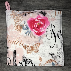 May include: Square, quilted pot holder with a pink loop. The design features a large pink rose, butterflies, the Eiffel Tower, and script on a white background. The word "Paris" is written in black script.
