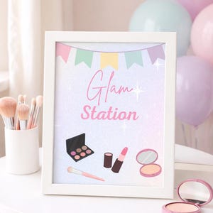May include: A framed print with the words "Glam Station" in pink script, featuring illustrations of makeup items like an eyeshadow palette, lipstick, and a compact mirror. Pastel balloons and makeup brushes are in the background.