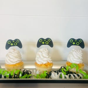 May include: Three cupcakes decorated with black and green video game controller toppers. The cupcakes are on a silver tray with black and green curling ribbon.