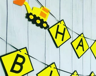 May include: A yellow and black construction-themed banner with the word "Happy" spelled out on individual yellow signs. Each sign has a black border and black letters. A yellow construction vehicle with black wheels is hanging from the banner.